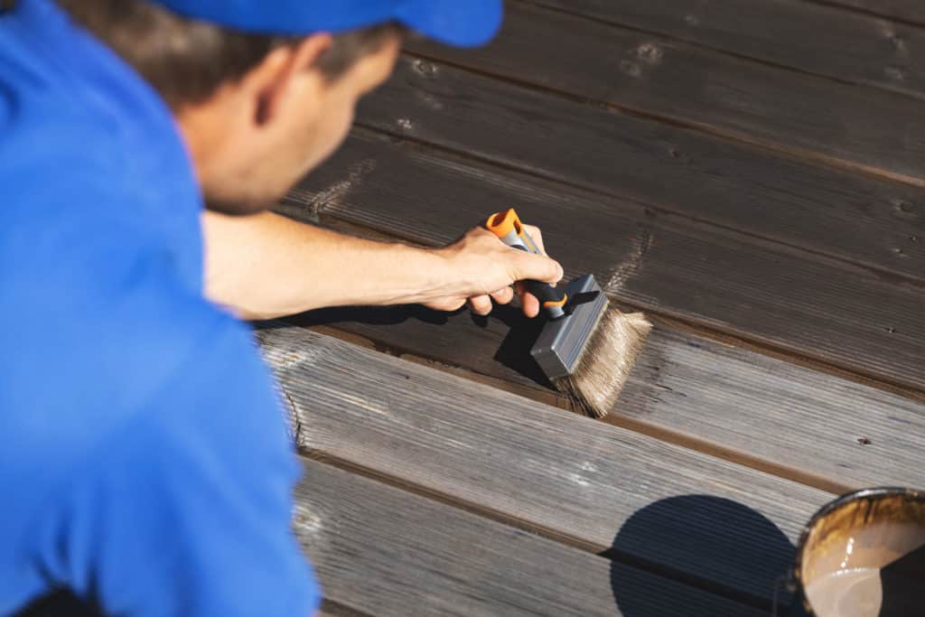 man in blue tee shirt and hat applying fresh stain to decking boards