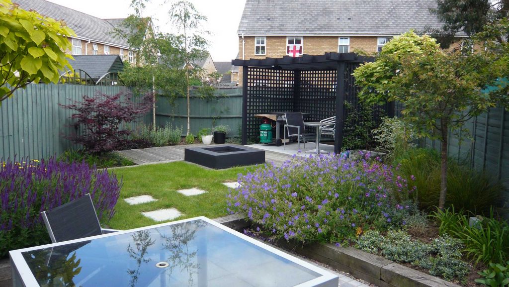 beautifully landscaped garden with dark timber pergola in far corner housing outdoor cooking equipment and garden furniture