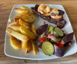 delicious meal on a white plate comprising steak topped with prawns, chips and a side salad