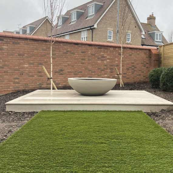 artificial grass walkway leading to decking and planter framed by two trees