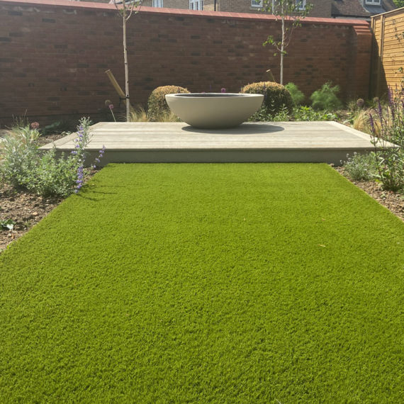 wide, artificial grass pathway leading to elegant planter placed on decking boards